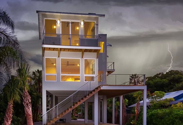 A multi-story gray home with white trim and large glass windows sits in a coastal setting with rain and lightning storms in the background. There are palm trees and other vegetation around the landscape.